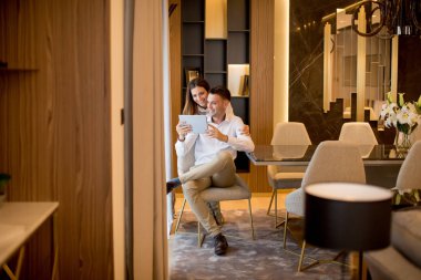 Genç çift evde dinleniyor ve pencerenin yanındaki oturma odasında dijital tablet kullanıyorlar.