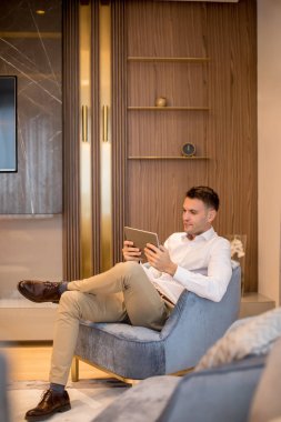 Yakışıklı genç adam lüks dairede oturuyor ve dijital tablet kullanıyor.