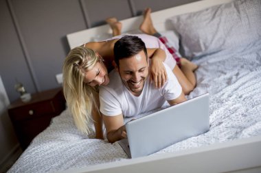 Intimate young lovers using laptop while lying on the bed in the bedroom