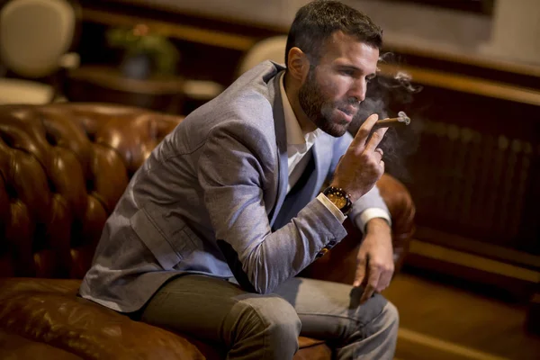 英俊的年轻人坐在皮革沙发上抽雪茄 — 图库照片