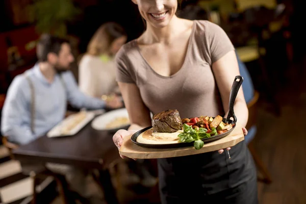 レストランで牛ステーキ付きの木製プレートを保持するかわいい若いウェイトレス — ストック写真