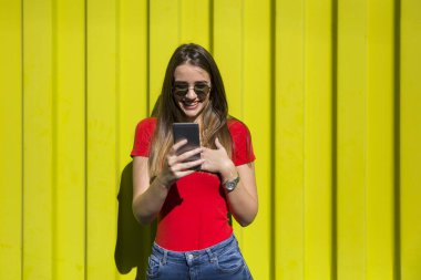 Sarı duvarın yanında cep telefonu kullanan güneş gözlüklü güzel bir kadın.