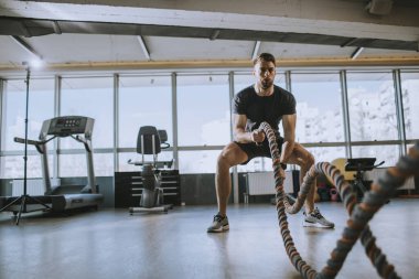 Yakışıklı genç adam spor salonunda savaş halatlarıyla pratik yapıyor.