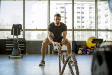 Yakışıklı genç adam spor salonunda savaş halatlarıyla pratik yapıyor.