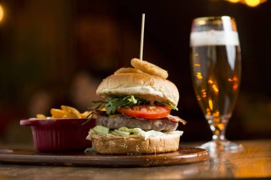 Eritilmiş çedar peynirli sığır burger, pastırma, domates, kızarmış soğanlı roket salatası ve masada baharatlı patates kızartması.