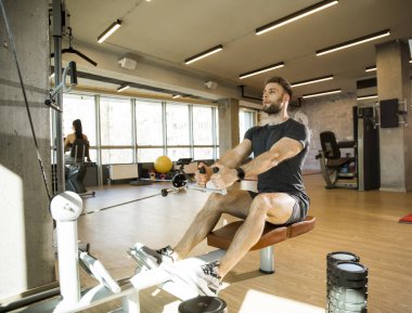 Yakışıklı genç adam spor salonunda oturan sıra makinesini kullanıyor.
