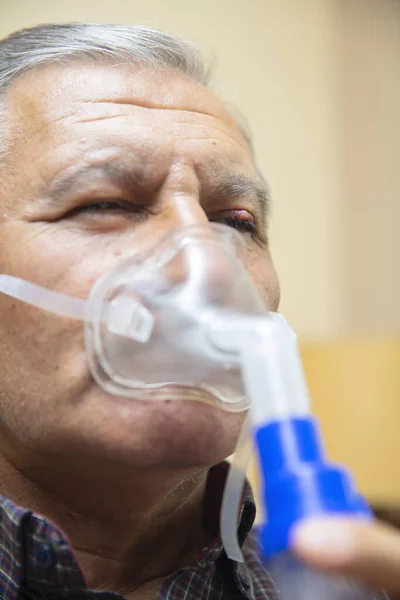 Senior man using medical equipment for inhalation with respiratory mask, nebulizer in the room
