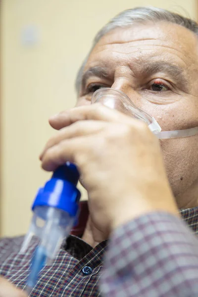 Senior man using medical equipment for inhalation with respiratory mask, nebulizer in the room