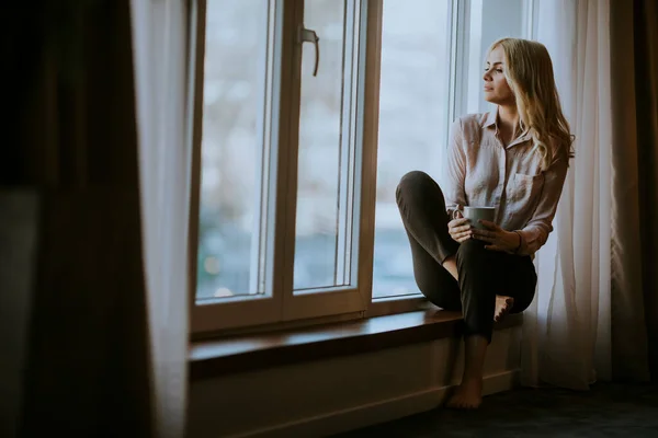 Pretty young woman with cup of tea or coffee sitting and drinking on the window sill at home