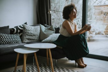 Güzel, genç, Afro-Amerikan, kıvırcık saçlı, pencerenin kenarında oturan ve elinde bir fincan kahve tutan genç bir kadın.