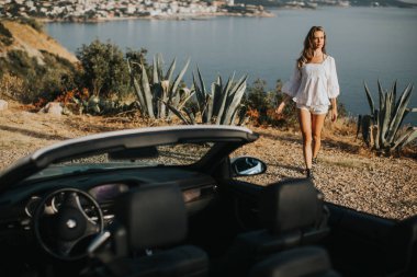 Cabriolet 'in deniz kenarındaki arabasına doğru yürüyen güzel genç bir kadın.