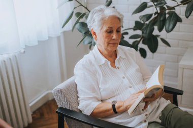 Evdeki odada kağıt kitap okuyan kıdemli bir kadın.