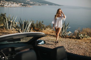 Cabriolet 'in deniz kenarındaki arabasına doğru yürüyen güzel genç bir kadın.