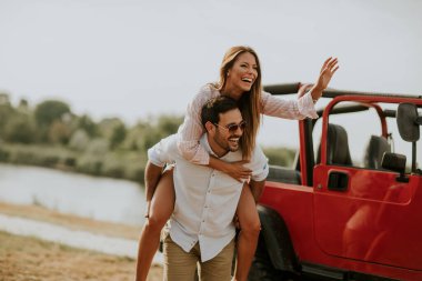 Genç bir kadın ve erkek yaz günü üstü açık kırmızı arabanın yanında eğleniyorlar.