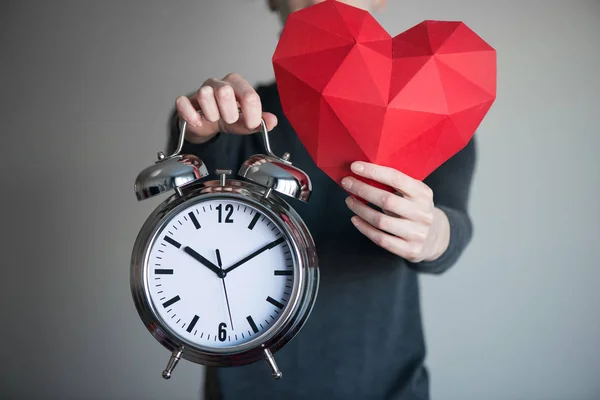 Wanita yang menunjukkan bentuk jantung poligonal merah dan jam alarm — Stok Foto