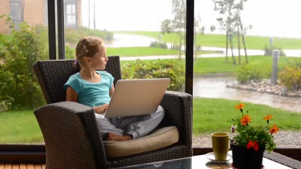 Маленькая девочка сидит на террасе в кресле с ноутбуком — стоковое видео