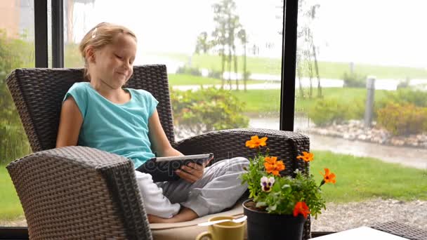 Niña sentada en la terraza en sillón con tablet digital — Vídeos de Stock