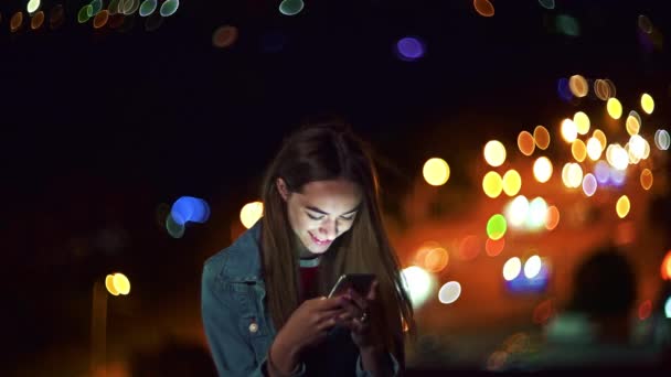 美しいですティーン女の子オン夜の街並みの背景とともに街路灯Bokeh保持携帯電話で手チャット — ストック動画