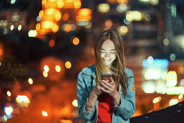 Teen girl on night cityscape background holding a phone in hand chatting — Stock Photo, Image