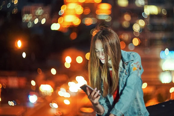 Dospívající dívka na noc cityscape pozadí drží telefon v ruce chatování — Stock fotografie