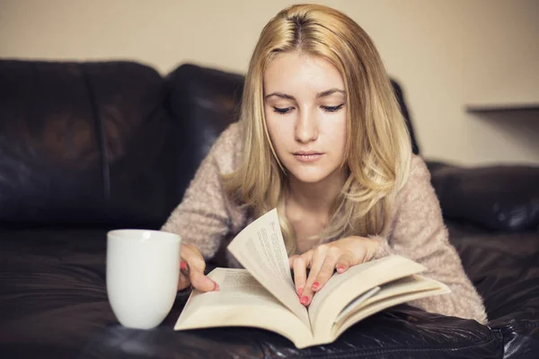 Молода жінка вдома, сидячи біля вікна, відпочиває у своїй вітальні читаючи книгу і п'ючи каву або чай — стокове фото