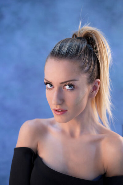 Красивая молодая блондинка с хвостиком, позирующая в студии перед синей стеной
