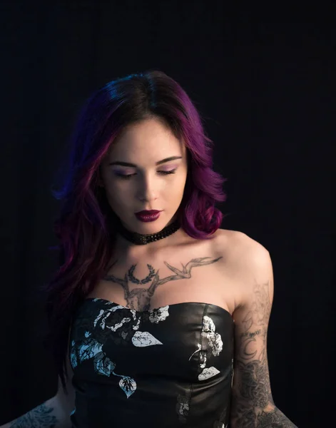 Beautiful girl with dyed hair and tattooes posing in studio before dark wall