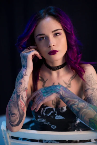 Beautiful girl with dyed hair and tattooes posing in studio before dark wall on chair