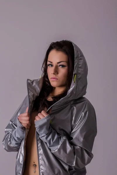 Beautiful young brunette woman in silver metalic coat on naked body posing in studio before light grey wall