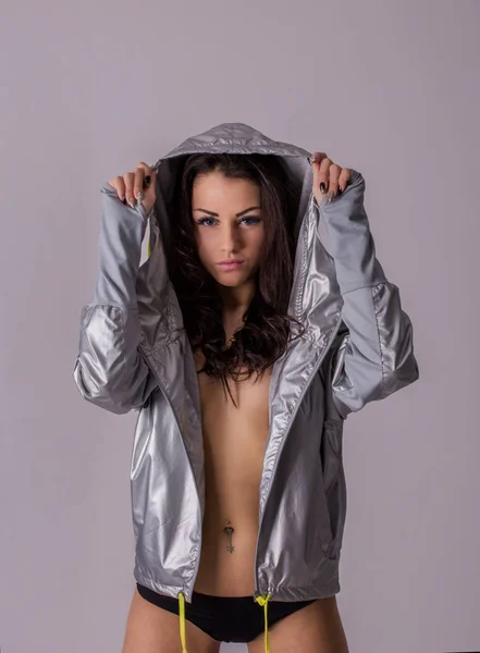 Beautiful young brunette woman in silver metalic coat on naked body posing in studio before light grey wall