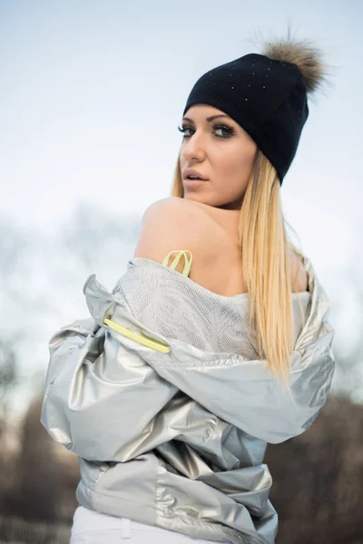 迷人的金发女人的肖像戴着帽子和银色夹克看着相机 而在模糊的冬季公园背景 — 图库照片
