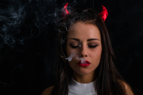 Young woman with Devil horns on head and cigarette smoke posing on black background