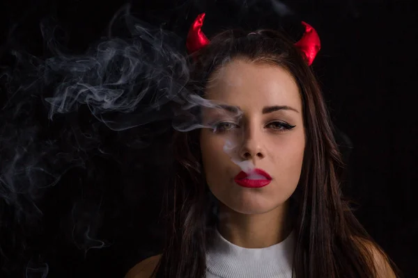 Young woman with Devil horns on head and cigarette smoke posing on black background