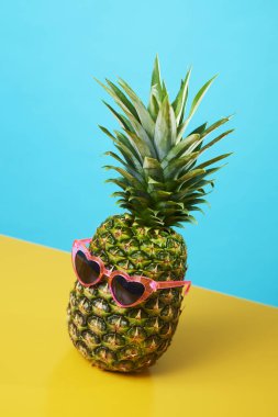 Mavi ve sarı arka planda pembe kalp şeklinde güneş gözlüğü takan bir ananas.
