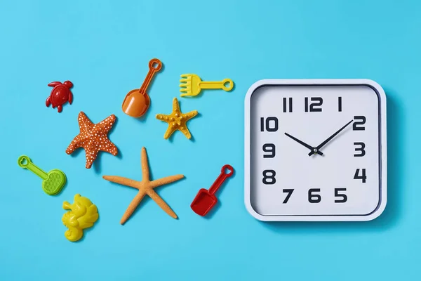 在蓝色的背景上 高角度的钟表 一些海星和一些不同的海滩玩具 如玩具铲子和球拍 以及一些不同颜色的砂模 — 图库照片