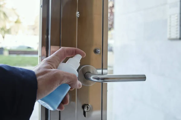 closeup of a caucasian man disinfecting the handle of the front door of an apartment building by spraying a blue sanitizer from a bottle