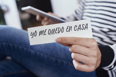 closeup of a man, in casual wear indoors, showing the message I stay at home in spanish written in a piece of paper, as a measure to stop the spreading of the covid-19 clipart