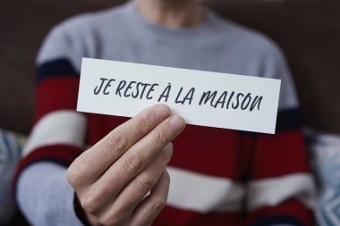 closeup of a man, in casual wear indoors, showing the message I stay at home in french written in a piece of paper, as a measure to stop the spreading of the covid-19 clipart
