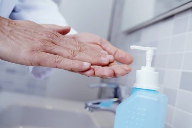 Beyaz önlüklü beyaz bir doktor ellerini mavi bir şişenin el dezenfektanı ile dezenfekte ediyor.