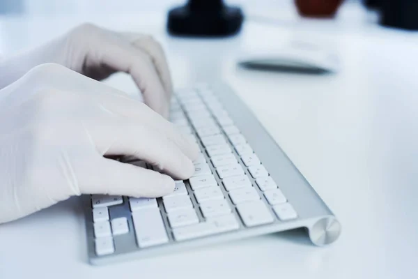 closeup of a man, wearing latex gloves, typing in a computer keyboard sitting at a white table