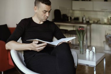 Kafkasyalı genç bir adama yakın çekim, günlük giysiler içinde, oturma odasında beyaz rahat bir sandalyede oturuyor, kitap okuyor.
