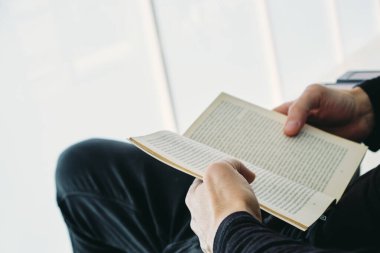 Kafkasyalı genç bir adamın yakın çekimi, günlük giysiler içinde, balkondaki açık bir koltukta oturmuş kitap okuyor.