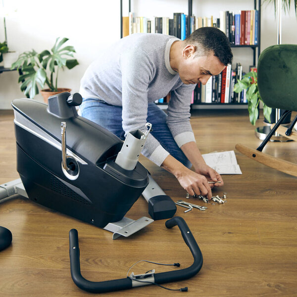 кавказский мужчина собирает различные части стационарного велосипеда в гостиной своего дома
