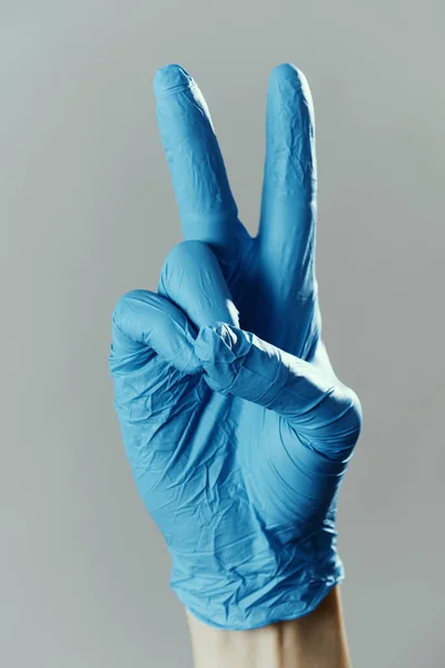 青い手術用手袋をした男の手を閉じて灰色の背景にVサインをした — ストック写真