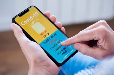 Kafkasyalı genç bir adamın yakın çekimi, rahat bir şekilde koltukta oturuyor, akıllı telefonundaki simülasyon uygulamasında covid-19 semptomlarını kontrol ediyor.