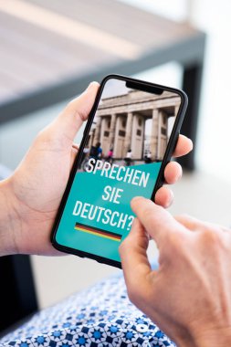 Kafkasyalı genç bir adamın yakın plan fotoğrafı, günlük kıyafetler giymiş, kapalı bir sandalyede oturuyor, akıllı telefonu elinde metinle ekranında Almanca yazıyor musunuz?