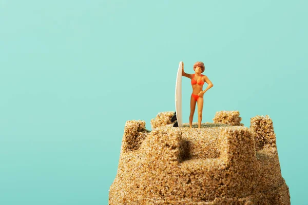 水着姿の小さな女性が砂の城の上のサーフボードの横に立って左側に空白がある青い背景に — ストック写真