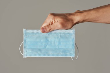 Gri bir arka planda, elinde cerrahi maske olan genç bir beyaz adamın yakın çekimi.