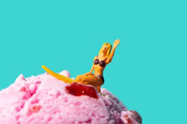 closeup of a miniature woman in swimsuit, kneeling on a surfboard, on a strawberry ice cream ball, against a blue background with some blank space clipart