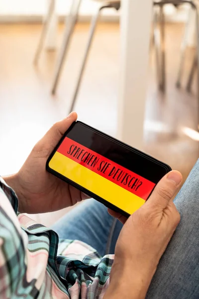 カジュアルな服を着た若い白人男性が椅子に座ってスマートフォンを手にテキストを持っているドイツ語で書かれたドイツ語をスクリーンで話す — ストック写真